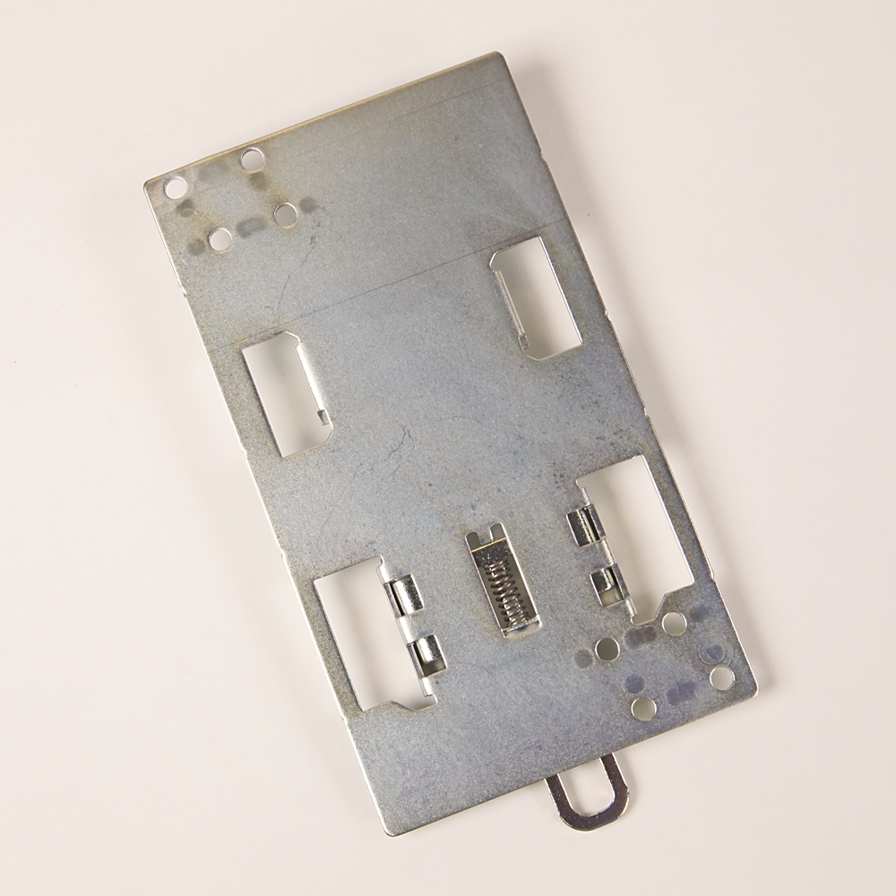 Metal mounting plate for an electronics module with several rectangular cutouts and a central spring-loaded contact.