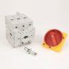 Three‑pole industrial circuit breaker with a red rotary handle on a yellow base and a metal mounting bolt in front.